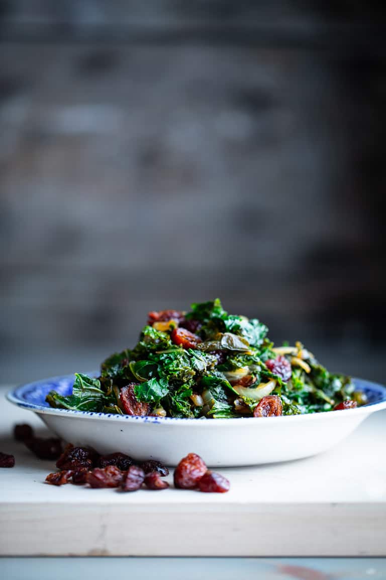 Side view of a bowl of kale with cranberries and balsamic vinegar on a gray table with a gray background