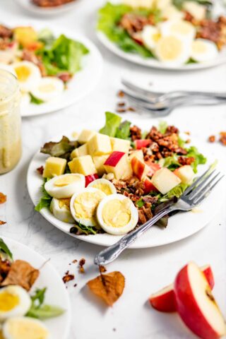 salads on a white table with apple slices, a jar of dressing and fall leaves