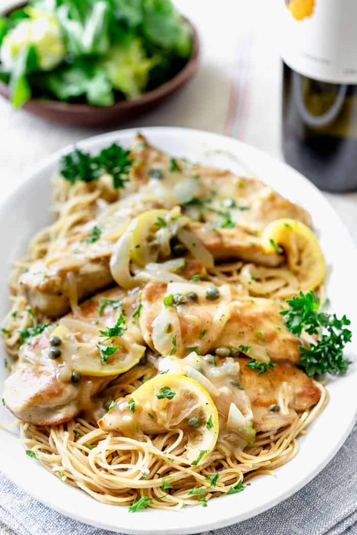 a white platter with chicken piccata, noodles and a bottle of wine