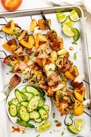 a tray of pork and peach kebabs with cucumber salad and limes