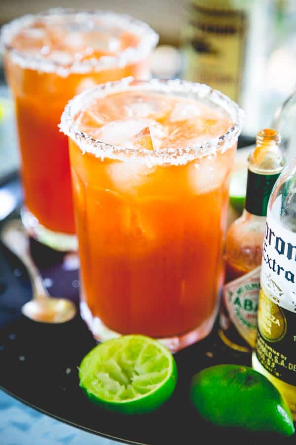 Two glasses of Michelada's on a black tray next to hot sauce, lime wedges and beer bottles