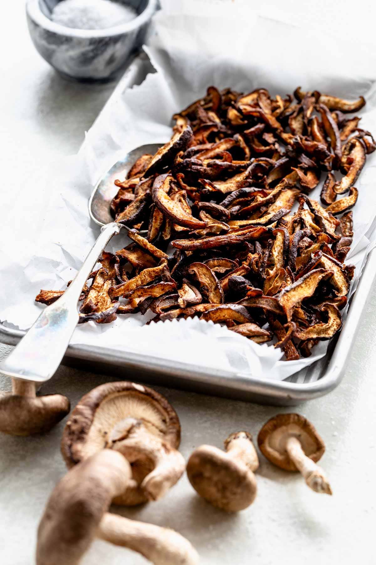 Shiitake mushroom bacon on baking parchment on a tray with a spoon on the tray, on a table. Raw shiitake mushrooms are next to the tray at the bottom of the picture.