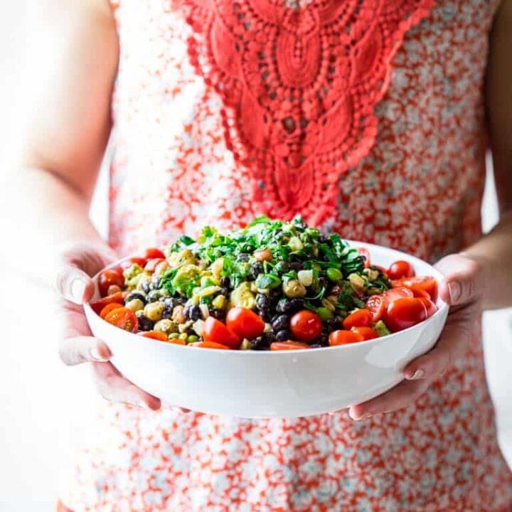 a woman's torso with a red shirt, hands holding a white bowl with a colorful bean salad with cherry tomatoes and cilantro