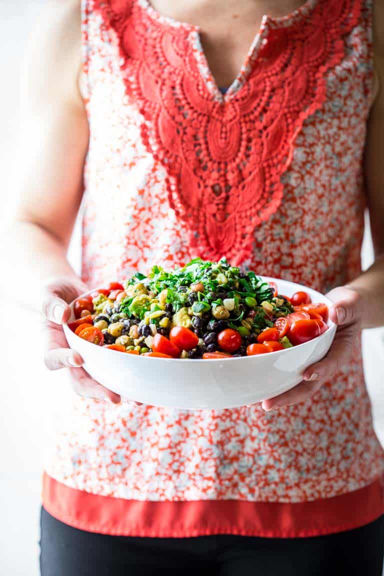 a woman's torso with a red shirt, hands holding a white bowl with a colorful bean salad with cherry tomatoes and cilantro