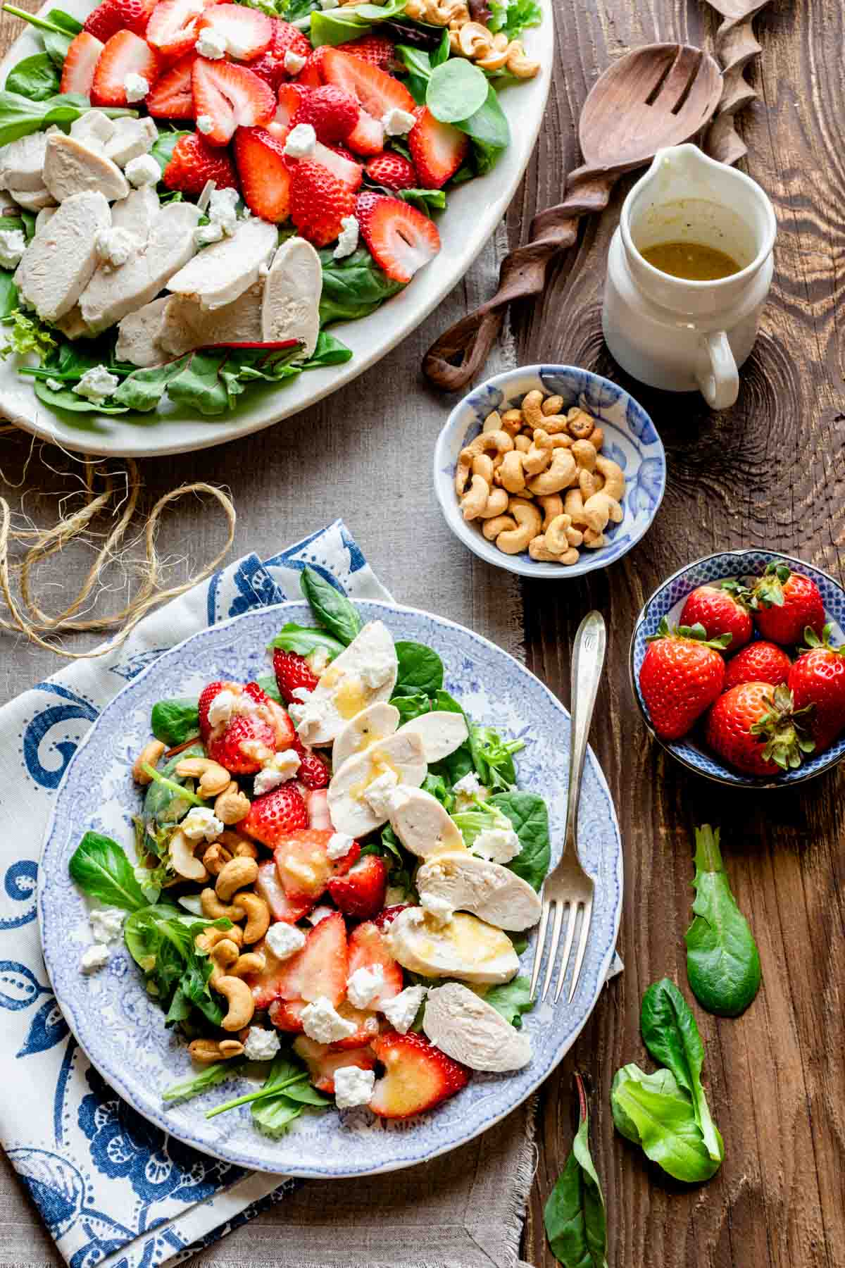 a platter with the salad comosed on it with a separate plate with a serving on it overhead on a table with pitcher of dressing and bowl of strawberries