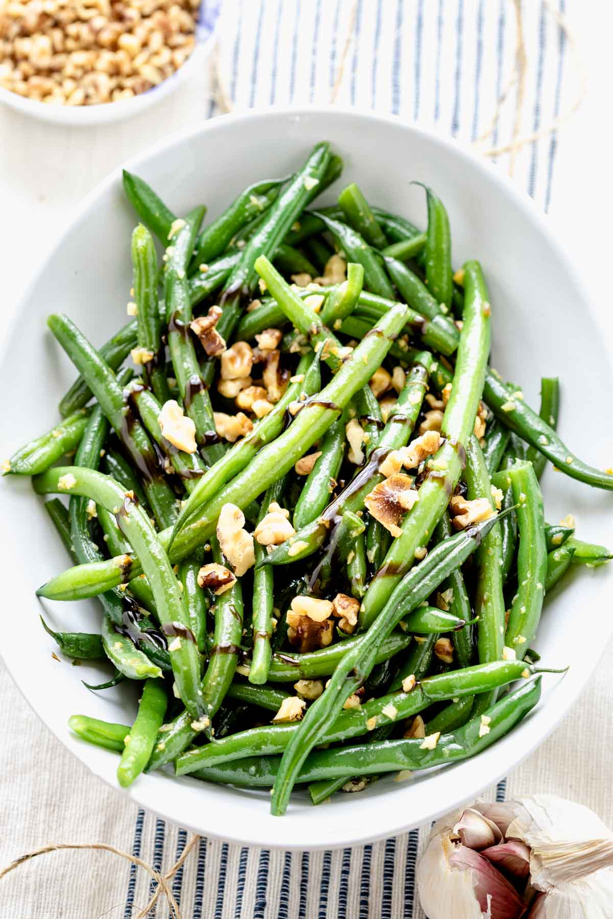 Green beans with balsamic and walnuts in a white serving bowl next to a small bowl of walnuts and a head of garlic