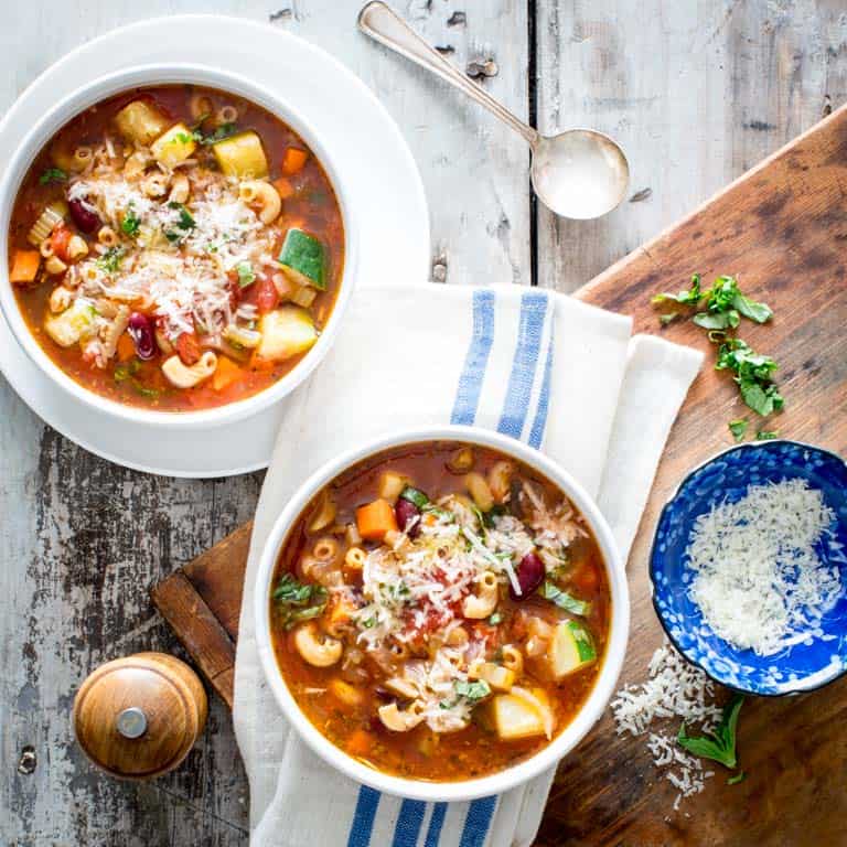 two white bowls of minestrone soup with a blue dish filed with grated Parmesan cheese
