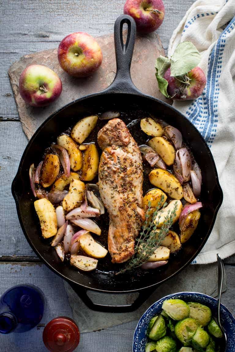A cast iron skillet with pork tenderloin on a gray painted surface