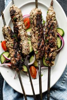four kofta kebabs on a platter with cucumbers and tomatoes