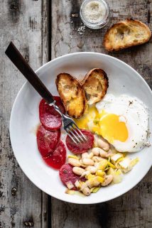 A white bowl with white beans and leeks, beets, a runny yolk egg and grilled baguette. With a wooden handled spoon resting on the dish.
