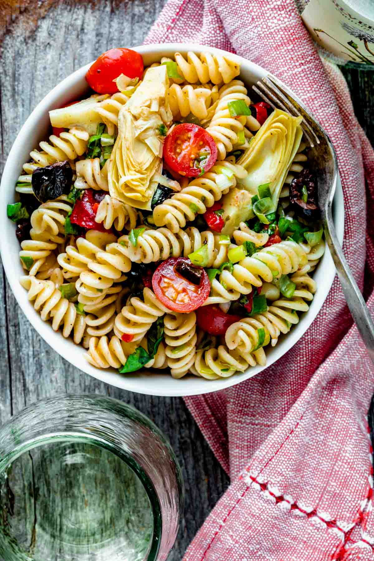 the pasta salad with artichoke hearts and cherry tomatoes in a small bowl on a red napkin
