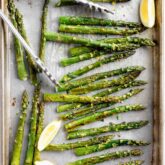 roasted asparagus on a sheet pan with tongs, herbs and lemon wedges