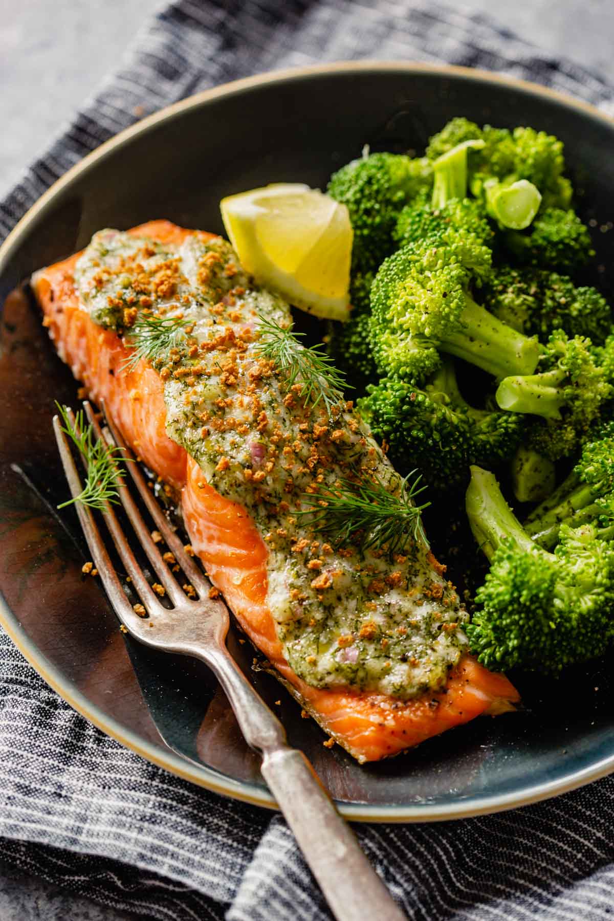 Salmon filet topped with creamy herb sauce set on a plate with broccoli, lemon wedge and a fork.