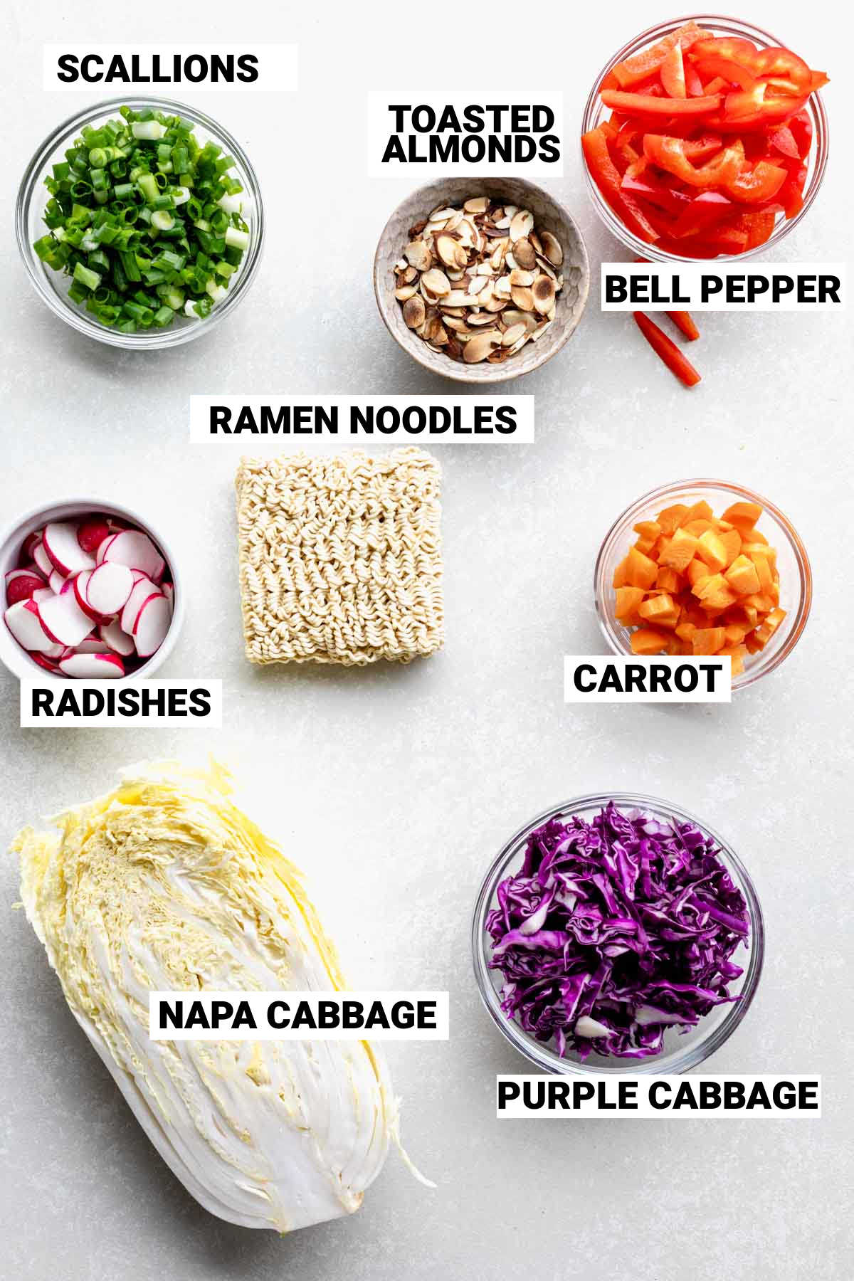 Vista superior de los ingredientes para la ensalada de fideos ramen de repollo Napa en tazones pequeños sobre una mesa con etiquetas de texto.