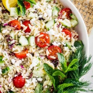 Close up of Orzo Pasta Salad in a white bowl on a table with a slice of lemon and a leafy garnish on top.
