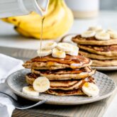 whole wheat banana pancakes stacked on a plate with banana slices and maple syrup drizzled on top
