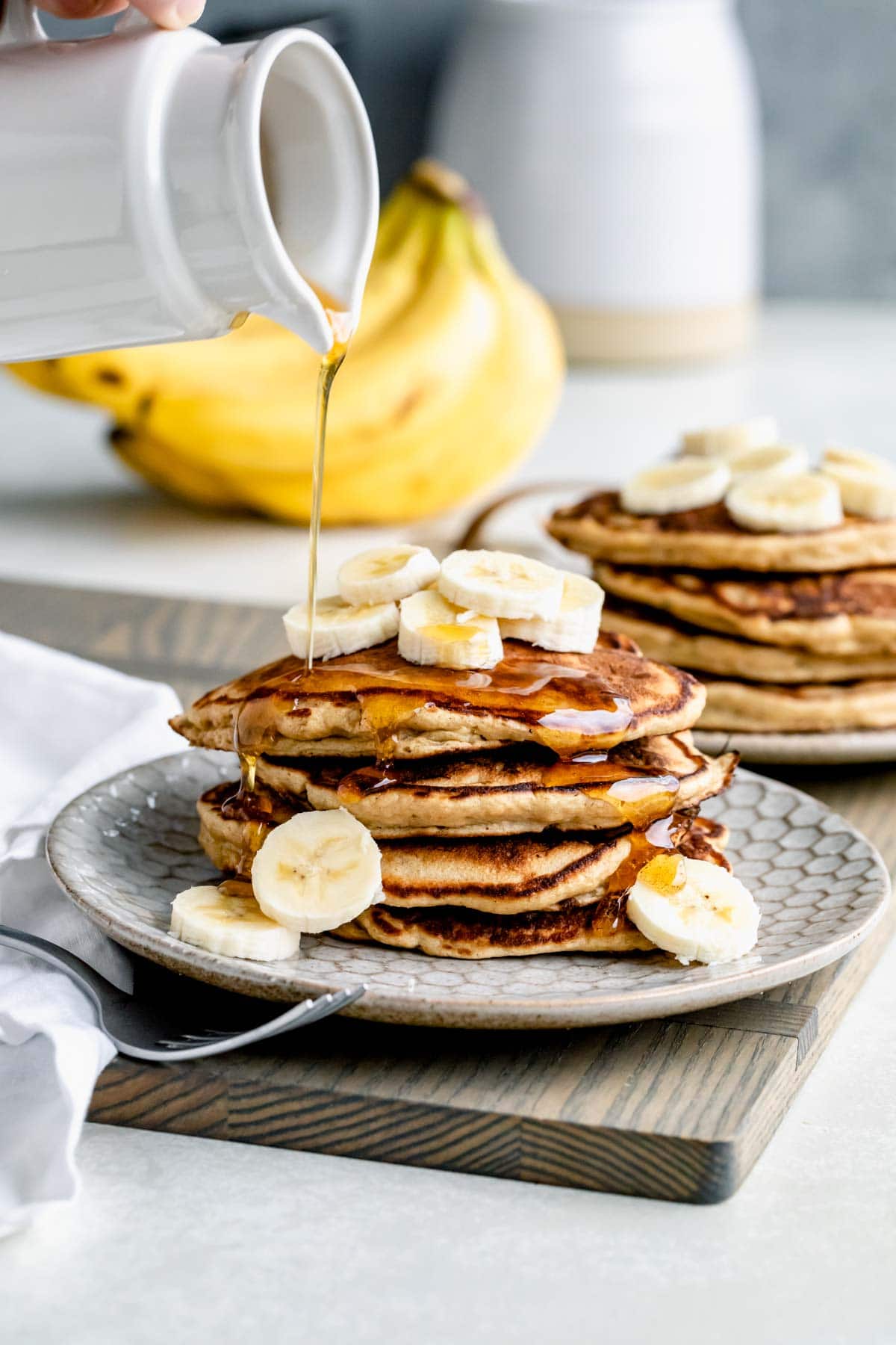 whole grain banana pancakes in a stack with sliced bananas on top and maple syrup drizzling from a carafe above