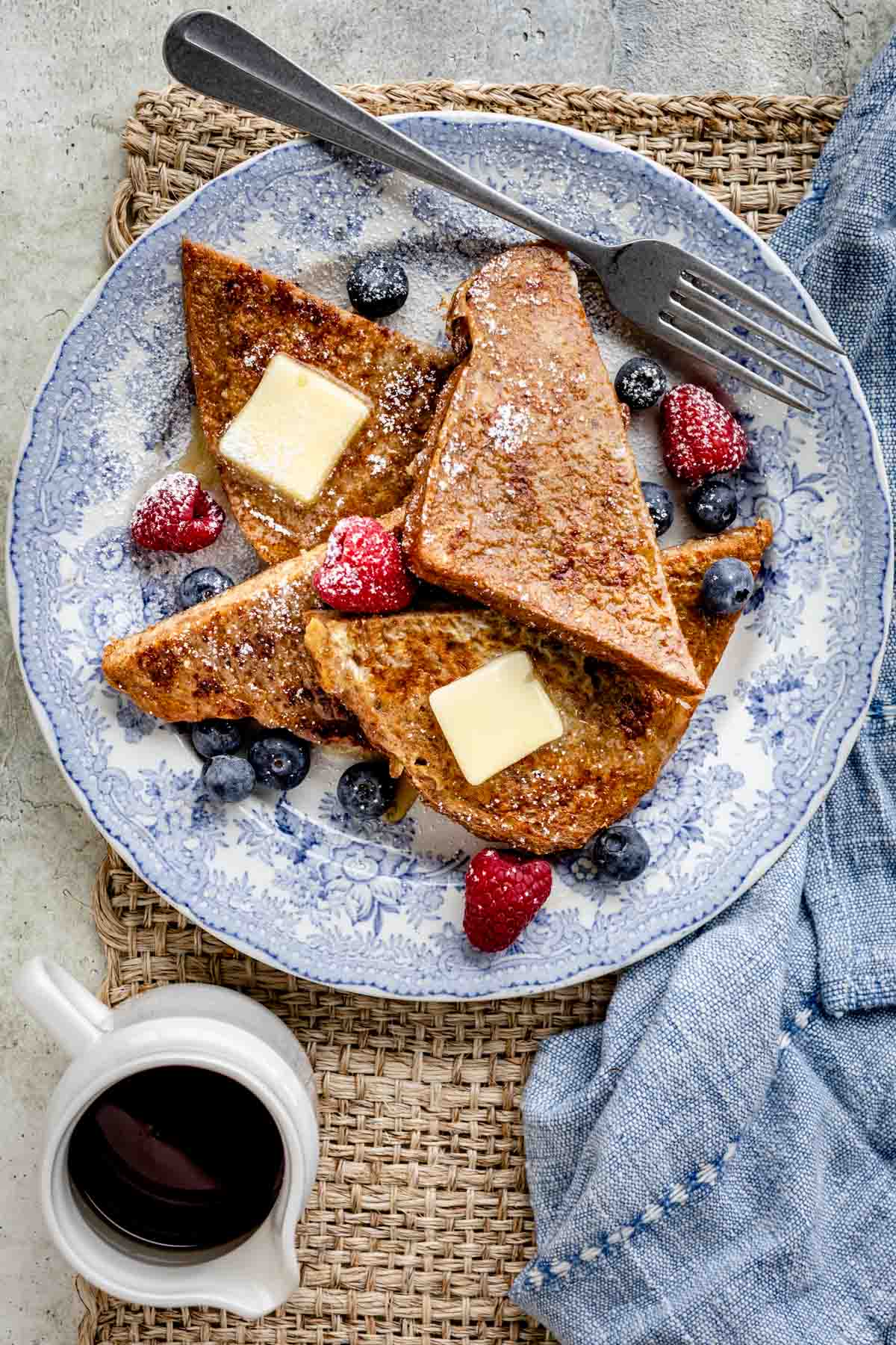 wheat bread french toast cut into triangles on a plate served with fruit, maple syrup, and pats of butter.
