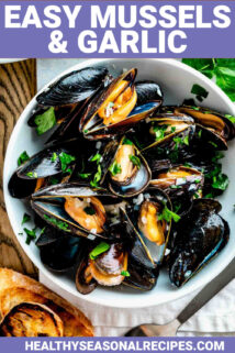 a white bowl filled with steamed mussels with parsley on top and two slices of grilled baguette on the table next to it
