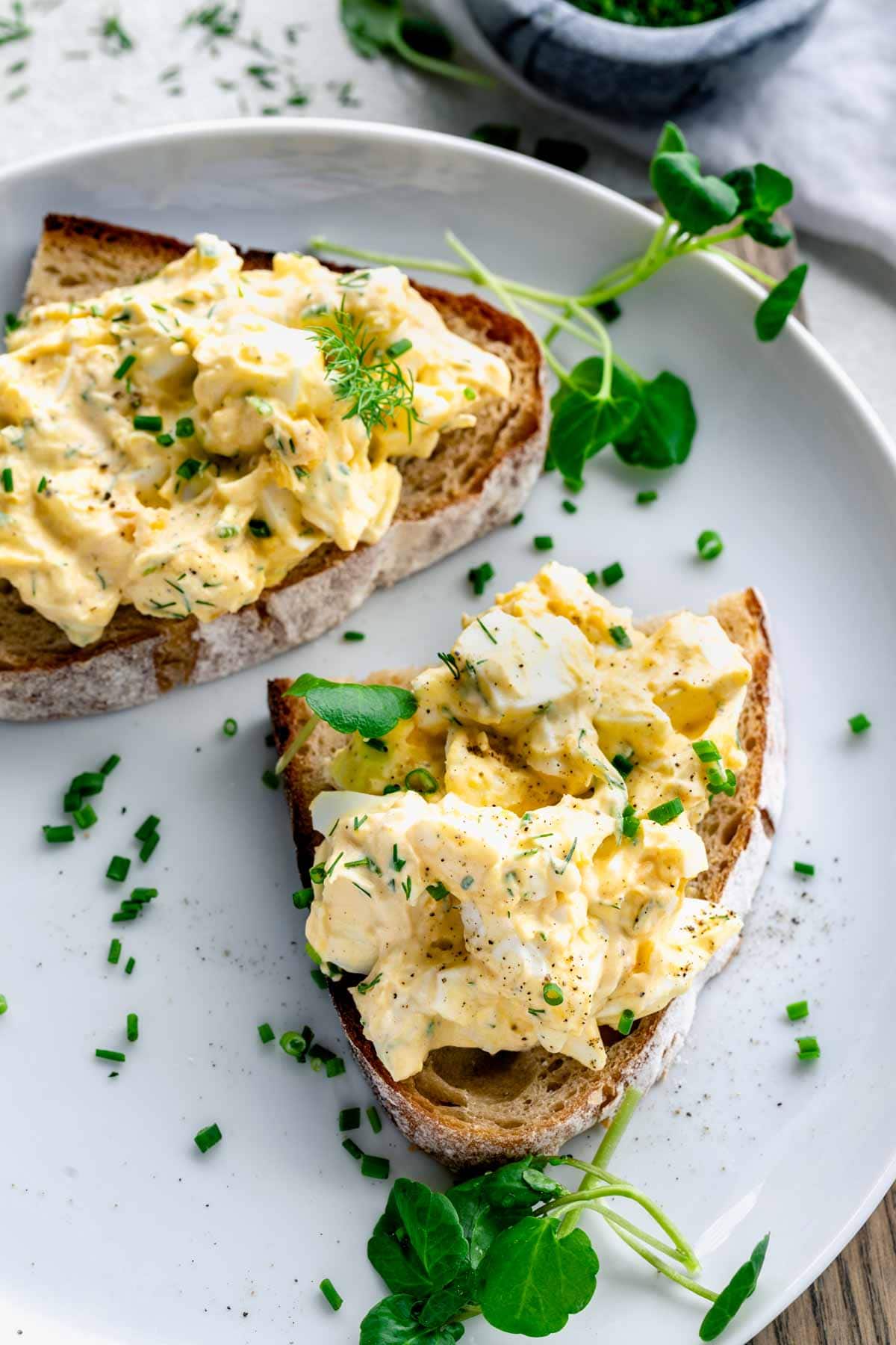 healthy egg salad on two slices of bread on a white plate with seasonings and garnish