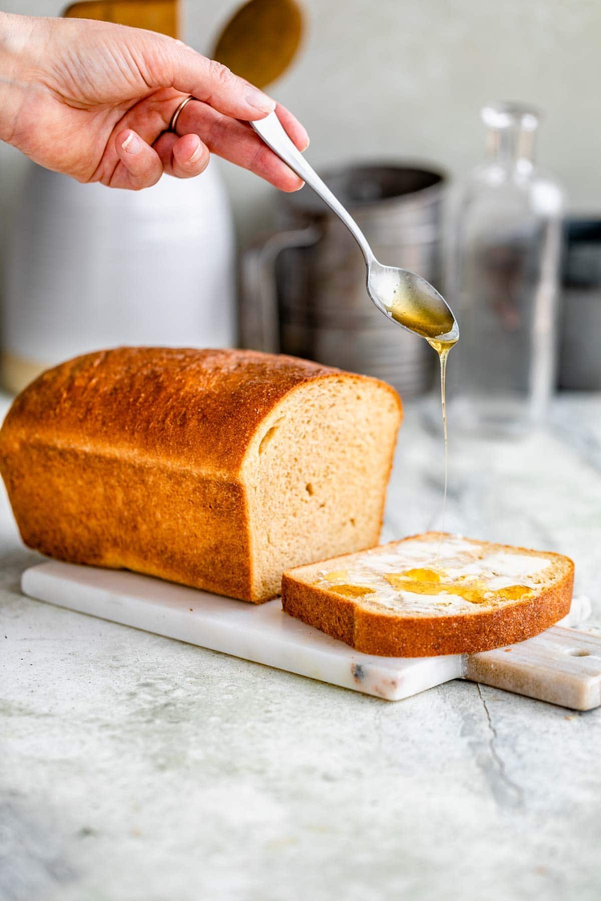 loaf of honey wheat bread with one slice cut off and butter smeared on it. A spoon is drizzling honey on top from above