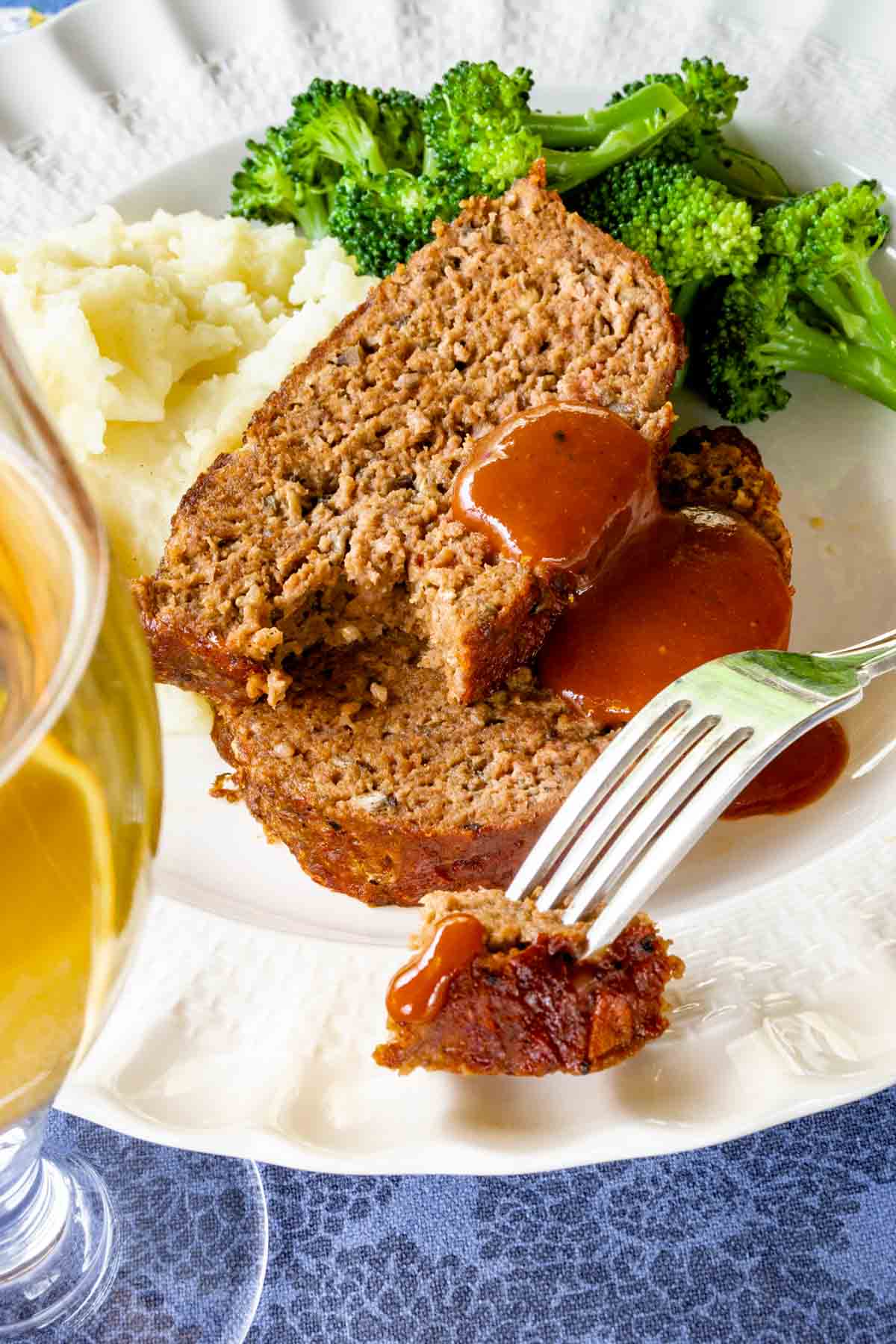 two slices of barbecued beef meatloaf with barbecue sauce on them, with mashed potatoes and broccoli