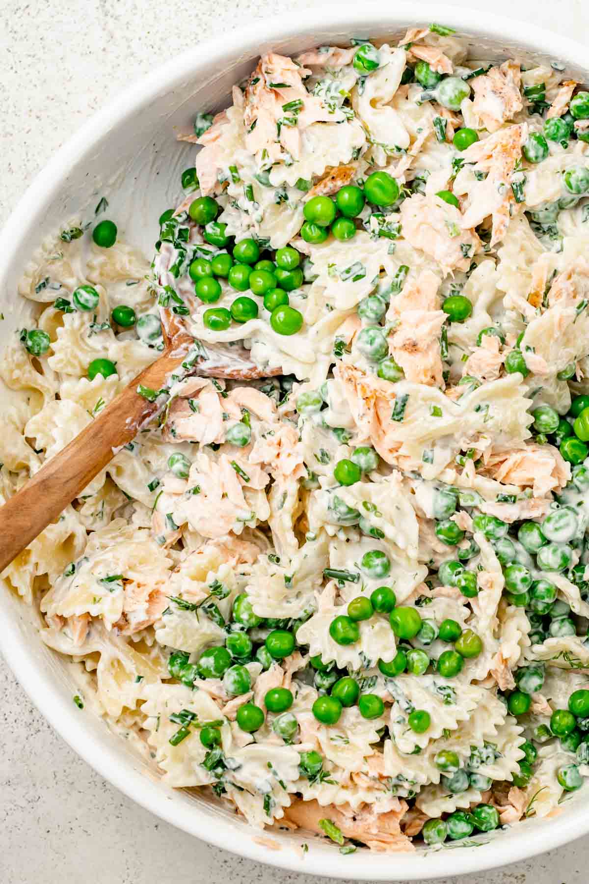 close up look at salmon pasta salad in a white bowl with extra peas on top and a wooden spoon on the side
