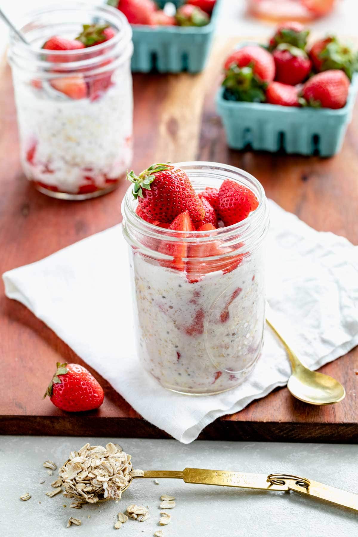 a focus on one mason jar full of overnight oats garnished with strawberries, with a gold spoon and strawberry in the foreground, and another mason jar of oats and some blue crates of strawberries in the background