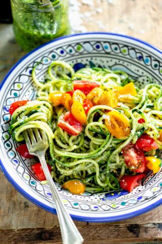 spiralized zucchini coated in pesto in a blue bowl with cherry tomatoes and a fork