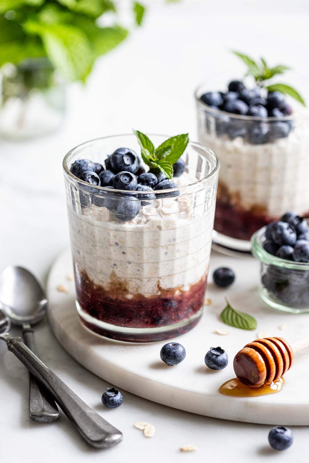 a couple of gl،es with mashed blueberries on the bottom, overnight oats layered on top, and fresh blueberries on the top. There is a clear bowl of blueberries to the right in the background, some s،s on the left, and ،ney leaking off of a ،ney s، in the foreground