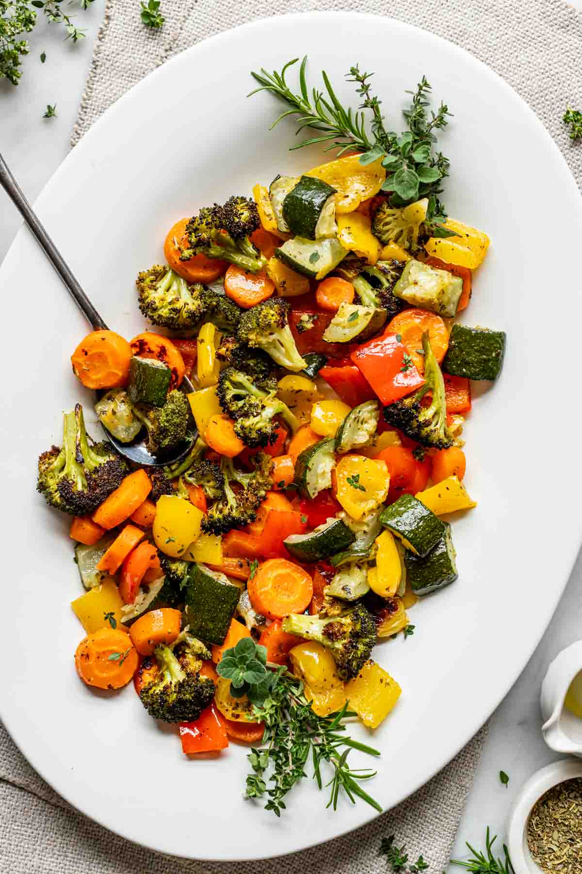 a large white serving bowl with a variety of sheet pan roasted vegetables in them, along with a serving spoon and fresh herbs for garnish.